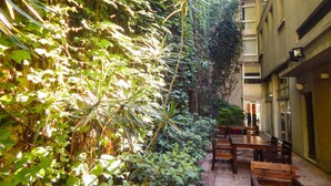 A narrow courtyard with a row of wooden tables and chairs, surrounded by dense foliage and a building with a balcony. - Ribera Sur Hotel (Buenos Aires)