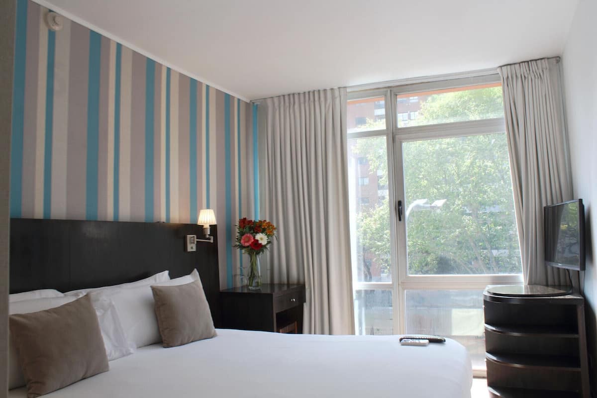 A hotel room with a bed, a nightstand with a lamp, a TV, and a window with curtains.