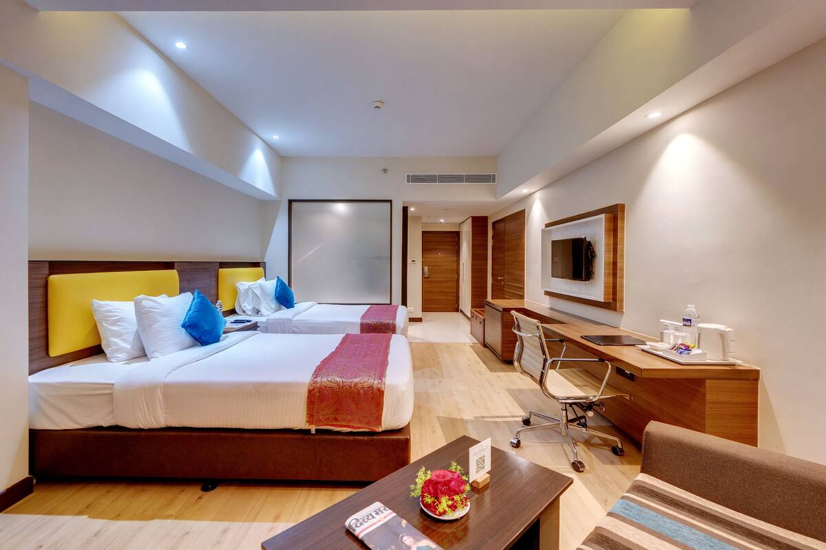 A hotel room with two beds, a desk, a chair, a TV, and a small table with a flower.