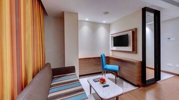 A modern hotel room with a sofa, a small table with a remote control, a blue chair, and a TV mounted on the wall.