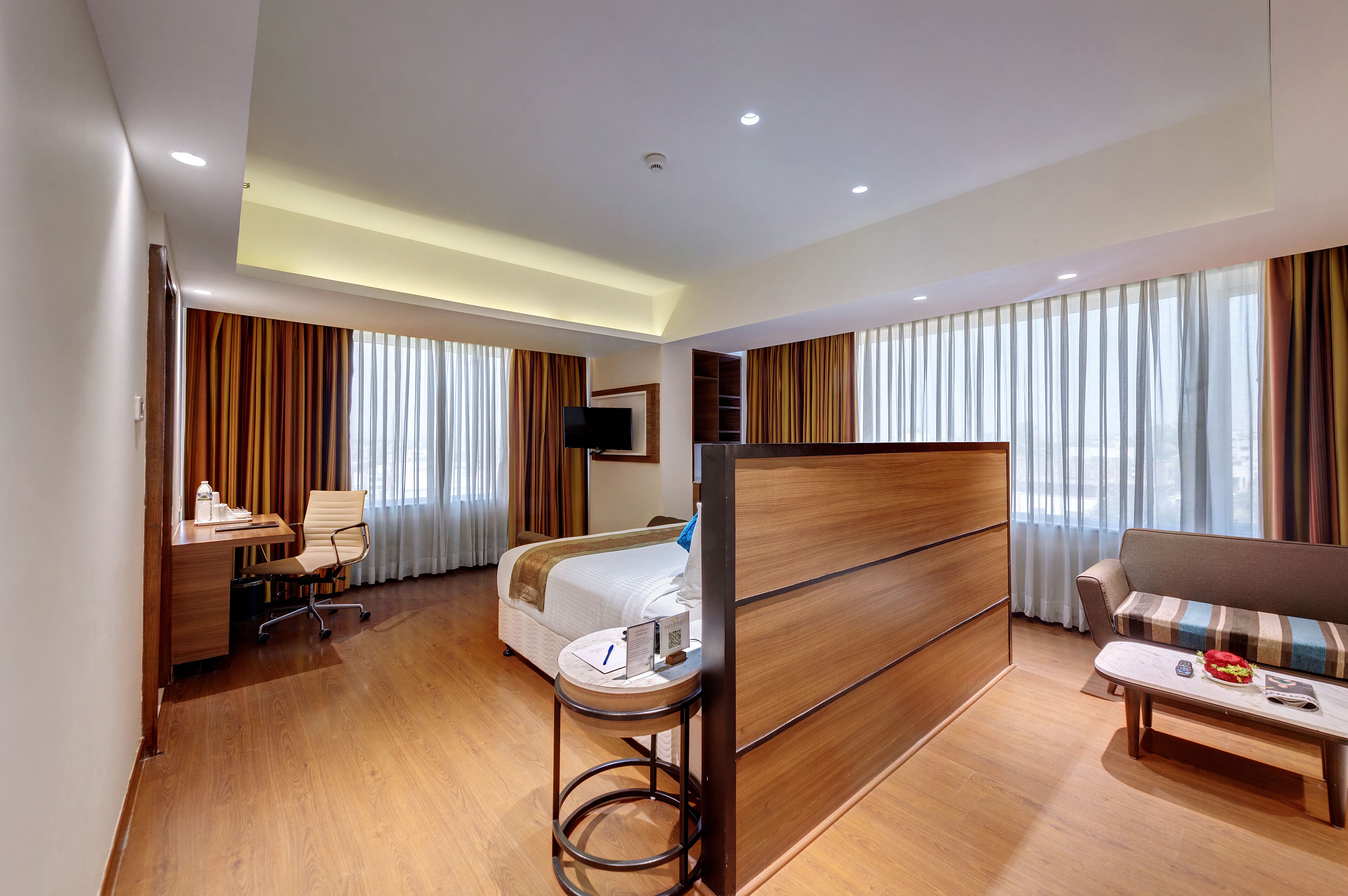 A modern hotel room with a wooden bed, a desk with a chair, a sofa, and a TV mounted on the wall.