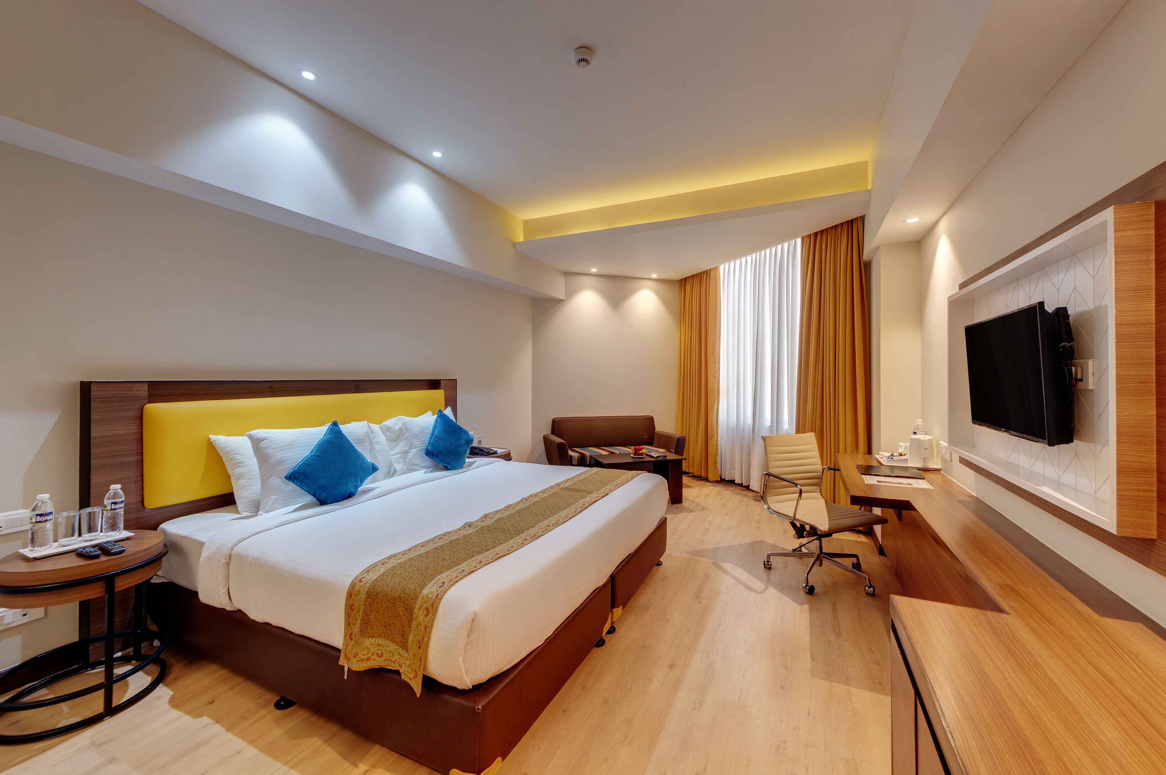 A hotel room with a large bed, a desk with a chair, a TV mounted on the wall, and a window with curtains.