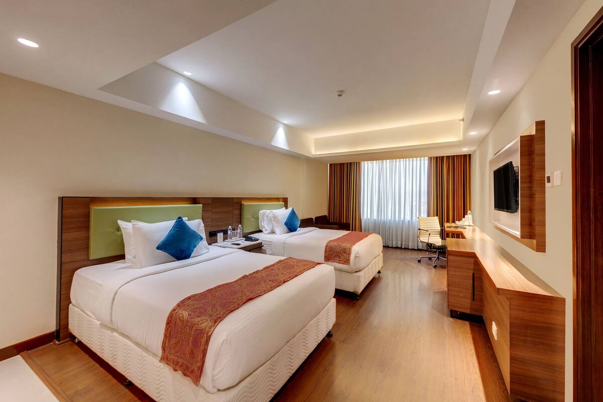 A hotel room with two beds, a desk, a chair, a TV, and a window with curtains.