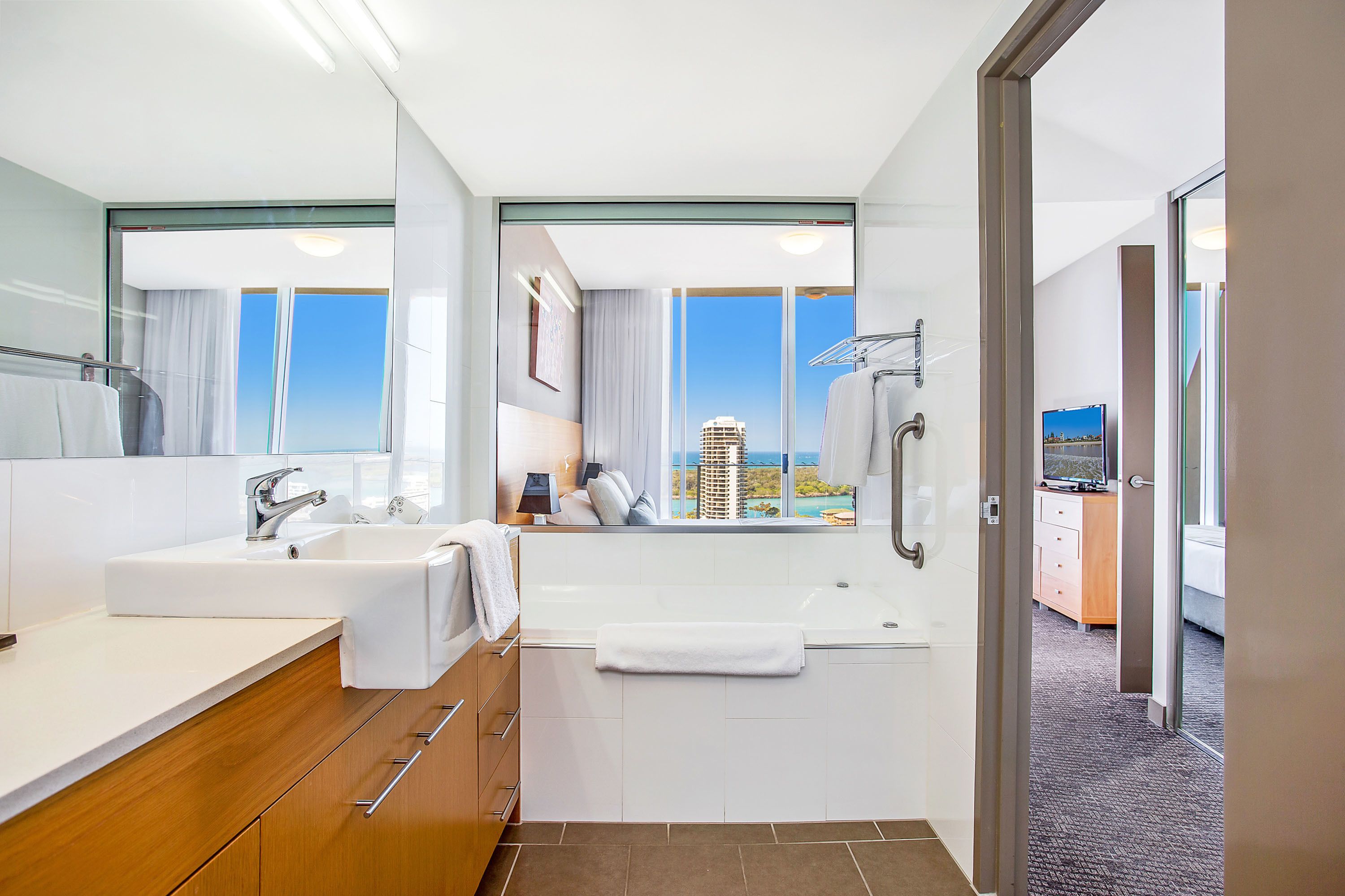 A modern bathroom with a large mirror, two sinks, a bathtub, and a view of a cityscape through large windows.