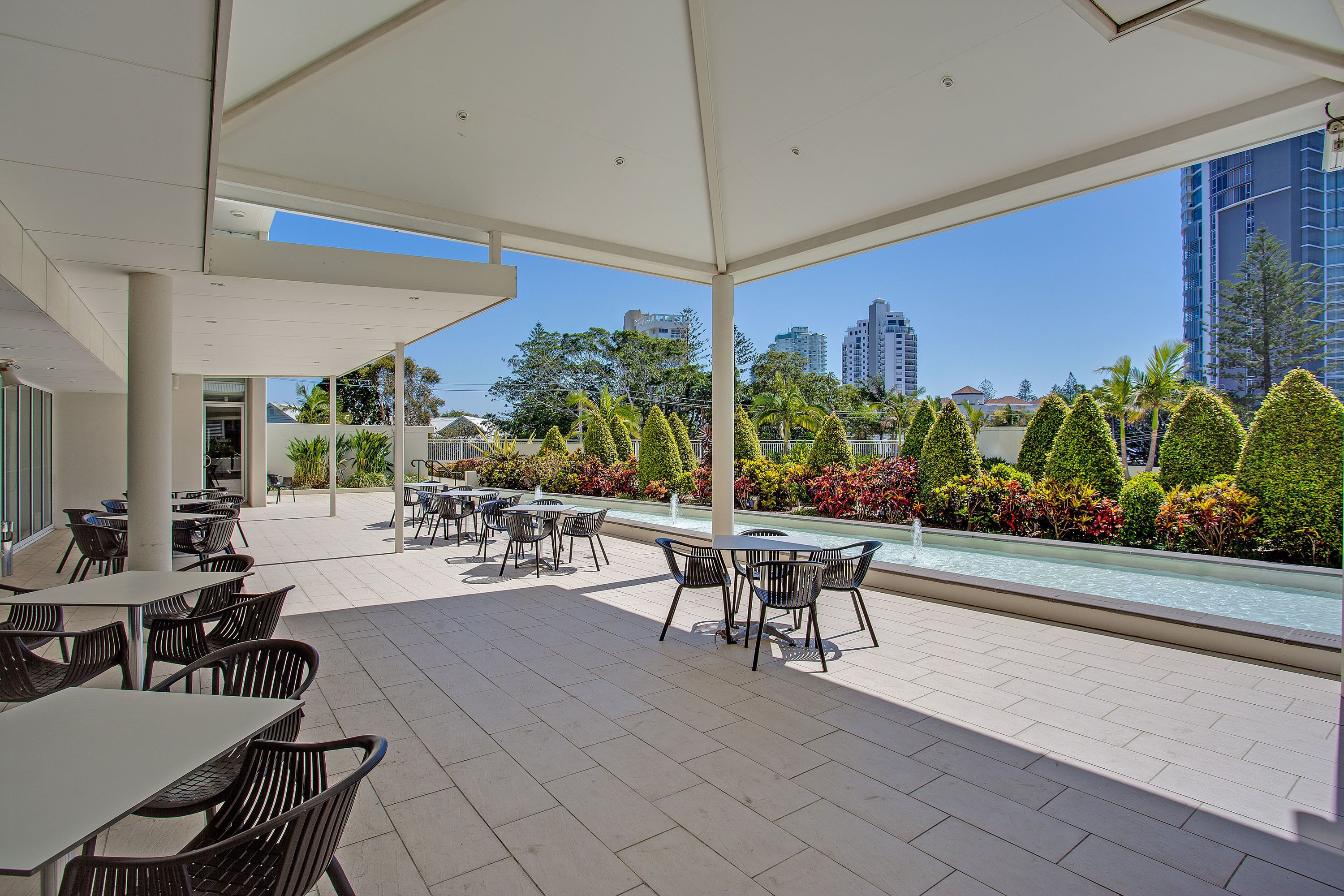 Un patio al aire libre con mesas y sillas, una piscina y vistas a edificios y árboles.