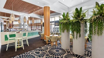 Eine moderne Hotelhalle mit Bar im Zentrum, Sitzbereichen und üppigen Topfpflanzen.