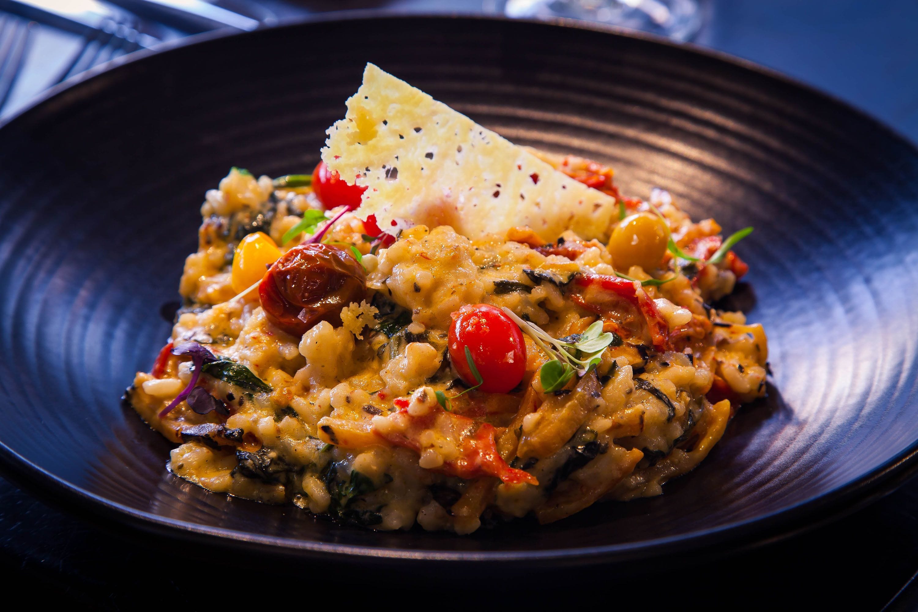 A plate of risotto with cherry tomatoes, herbs, and a piece of bread on a dark plate.
