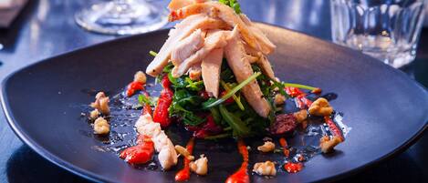 A black plate with a dish of sliced chicken, greens, and red peppers, accompanied by glasses of wine on a table.