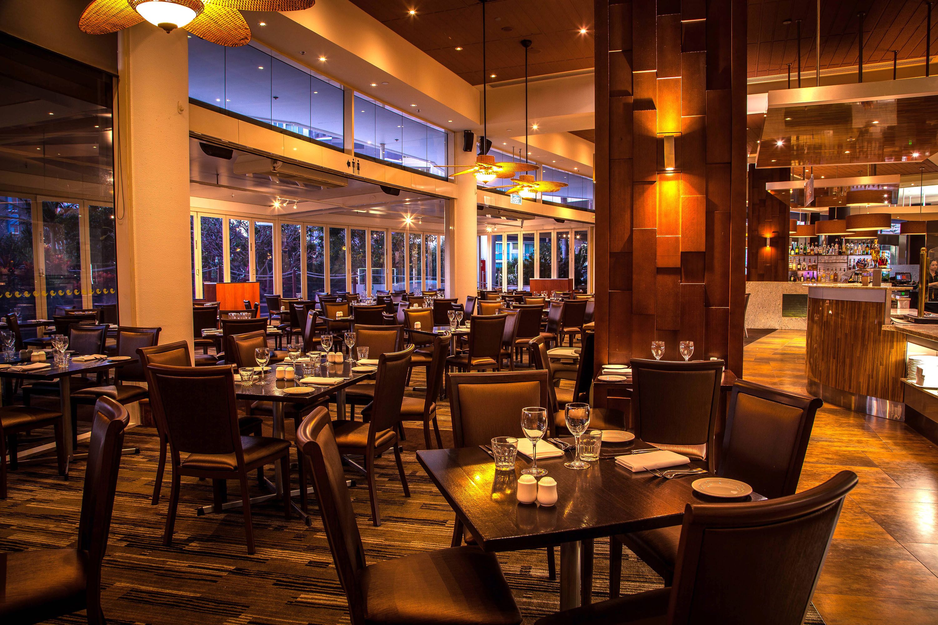 A spacious restaurant with multiple dining tables and chairs, large windows, and a bar area.