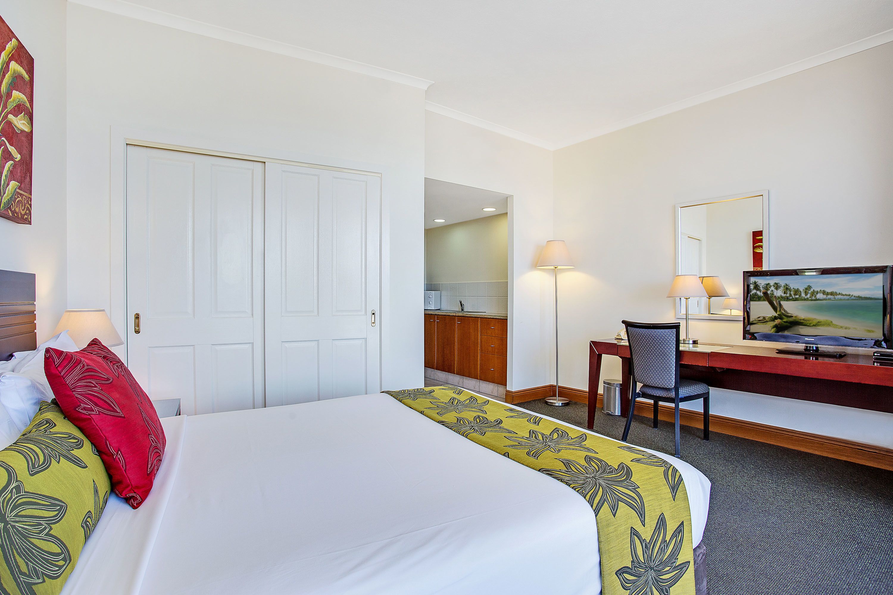 A hotel room with a bed, desk, chair, television, and a door leading to a kitchen area.