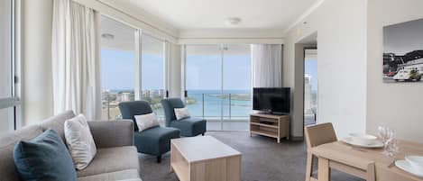 A modern hotel room with a balcony, a flat-screen TV, a dining area, and a large window offering a city view.