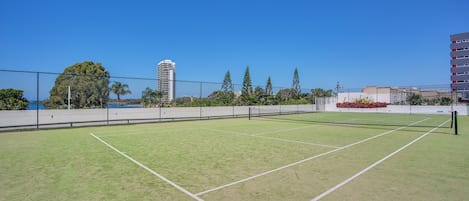 Una cancha de tenis con una red, rodeada por una valla, árboles y edificios al fondo.