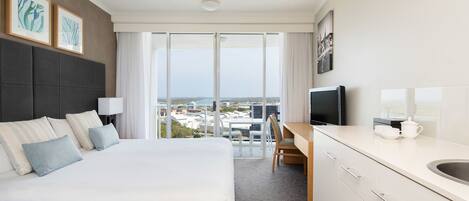 A hotel room with a large bed, a desk with a chair, a television, and a view of the city through large windows.