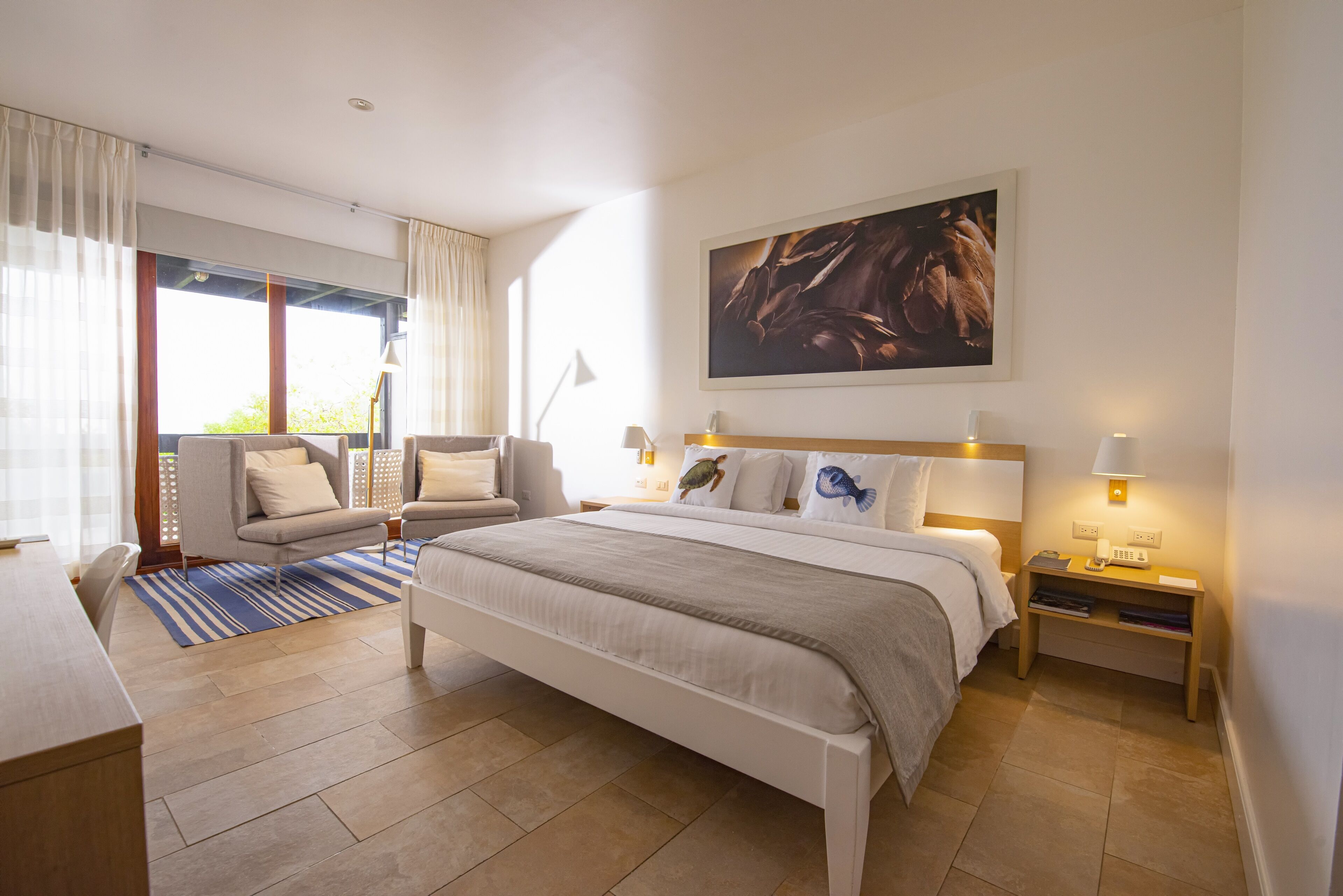 Quarto de hotel com cama, escrivaninha, cadeira, sofá, uma janela grande com cortinas e um quadro na parede.