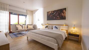 Habitación de hotel con cama, escritorio, silla, sofá, un ventanal con cortinas y un cuadro en la pared.