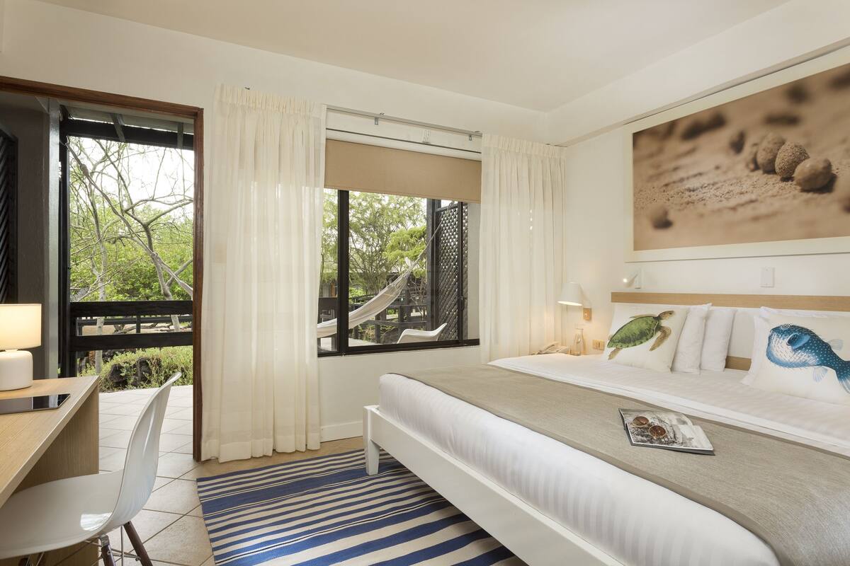 Una habitación de hotel con cama, escritorio, silla, ventana con vistas a árboles y un tobogán, y un cuadro en la pared.