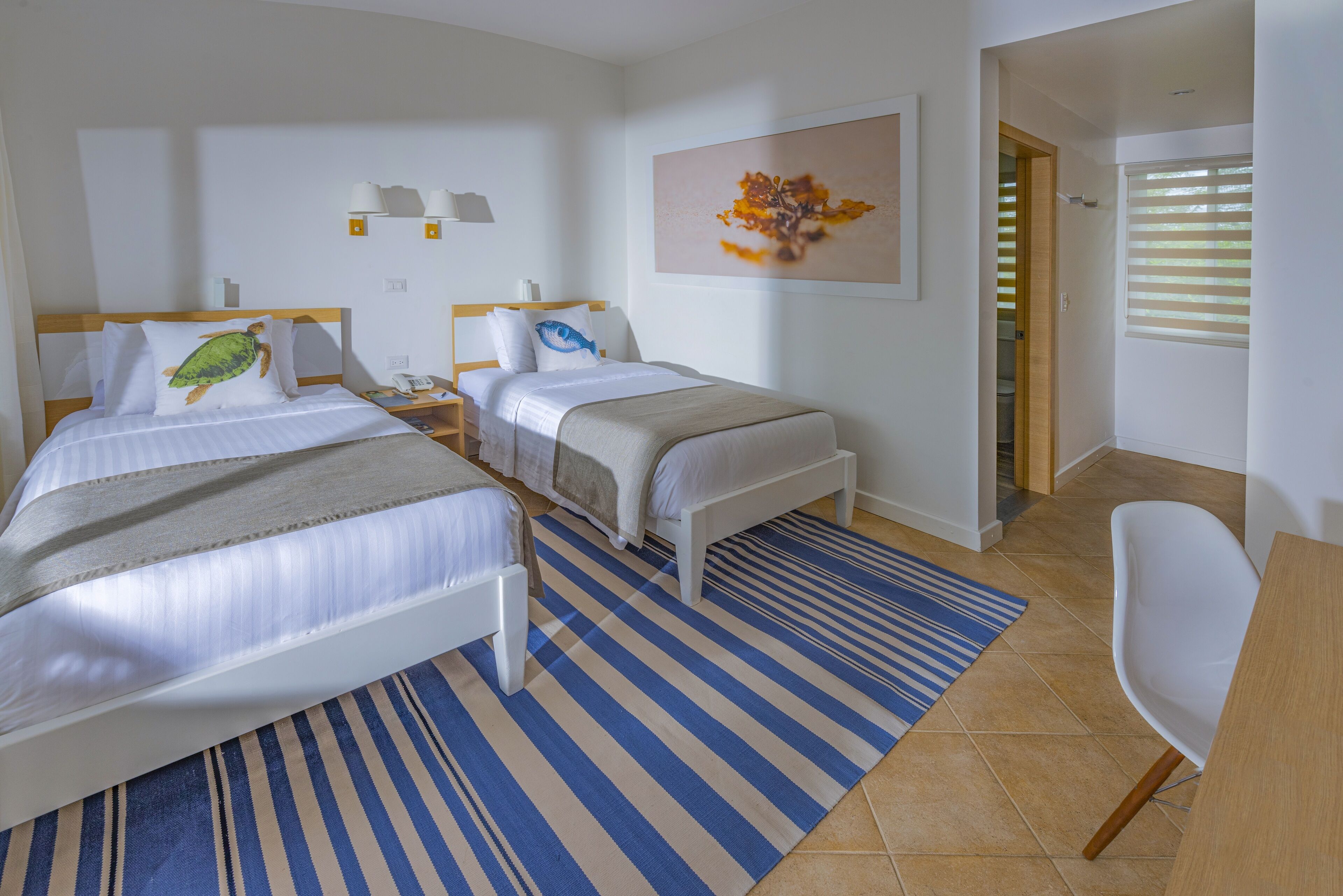 Une chambre d’hôtel avec deux lits, un bureau, une chaise, un tapis à rayures et un tableau au mur.