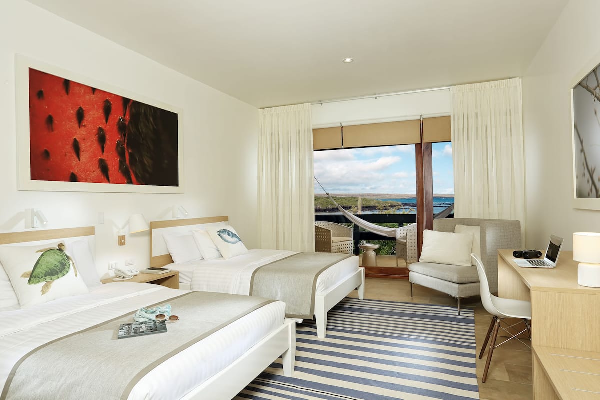 Habitación de hotel con dos camas, un escritorio, una silla, un ventanal con vistas y una hamaca afuera.