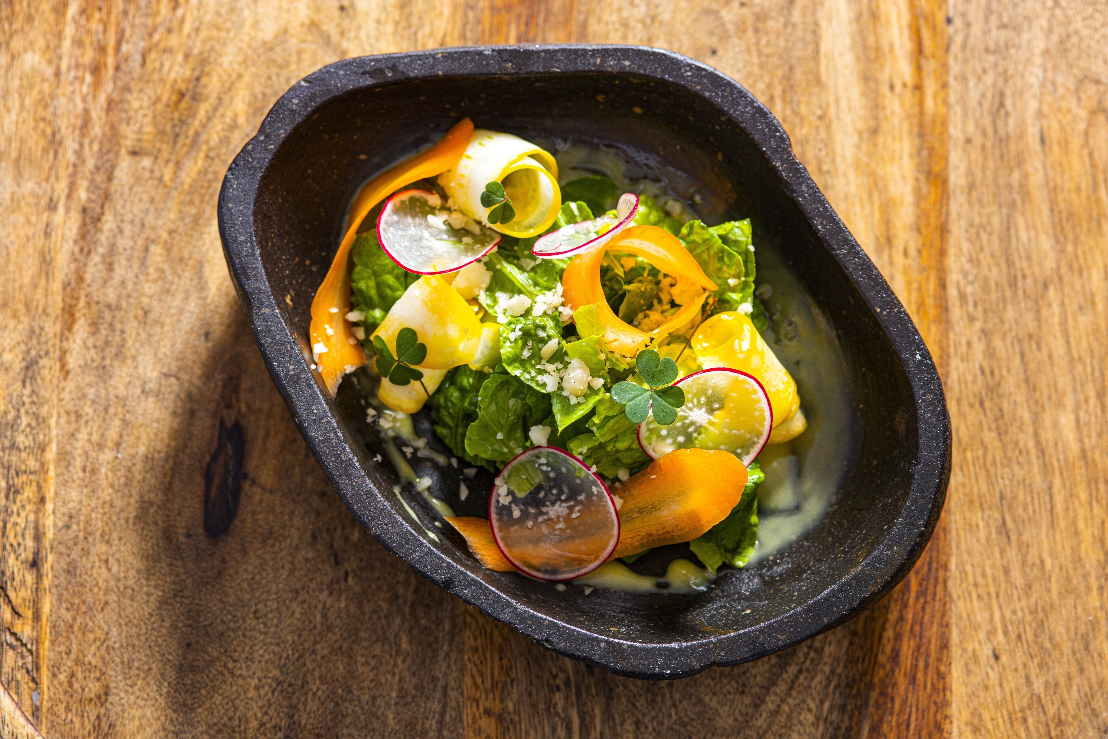 Un tazón de ensalada con diferentes verduras y queso sobre una superficie de madera.