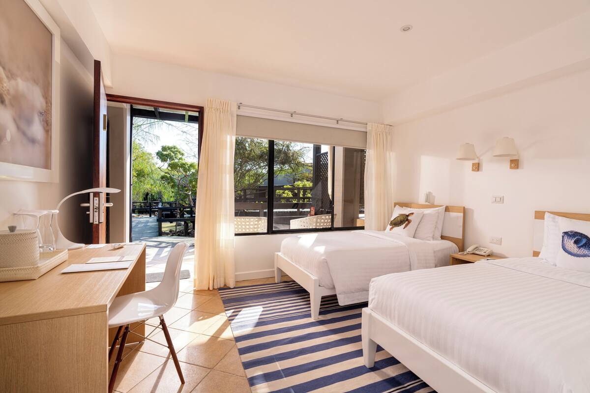 Una habitación de hotel con dos camas, un escritorio, una silla y un ventanal con cortinas.