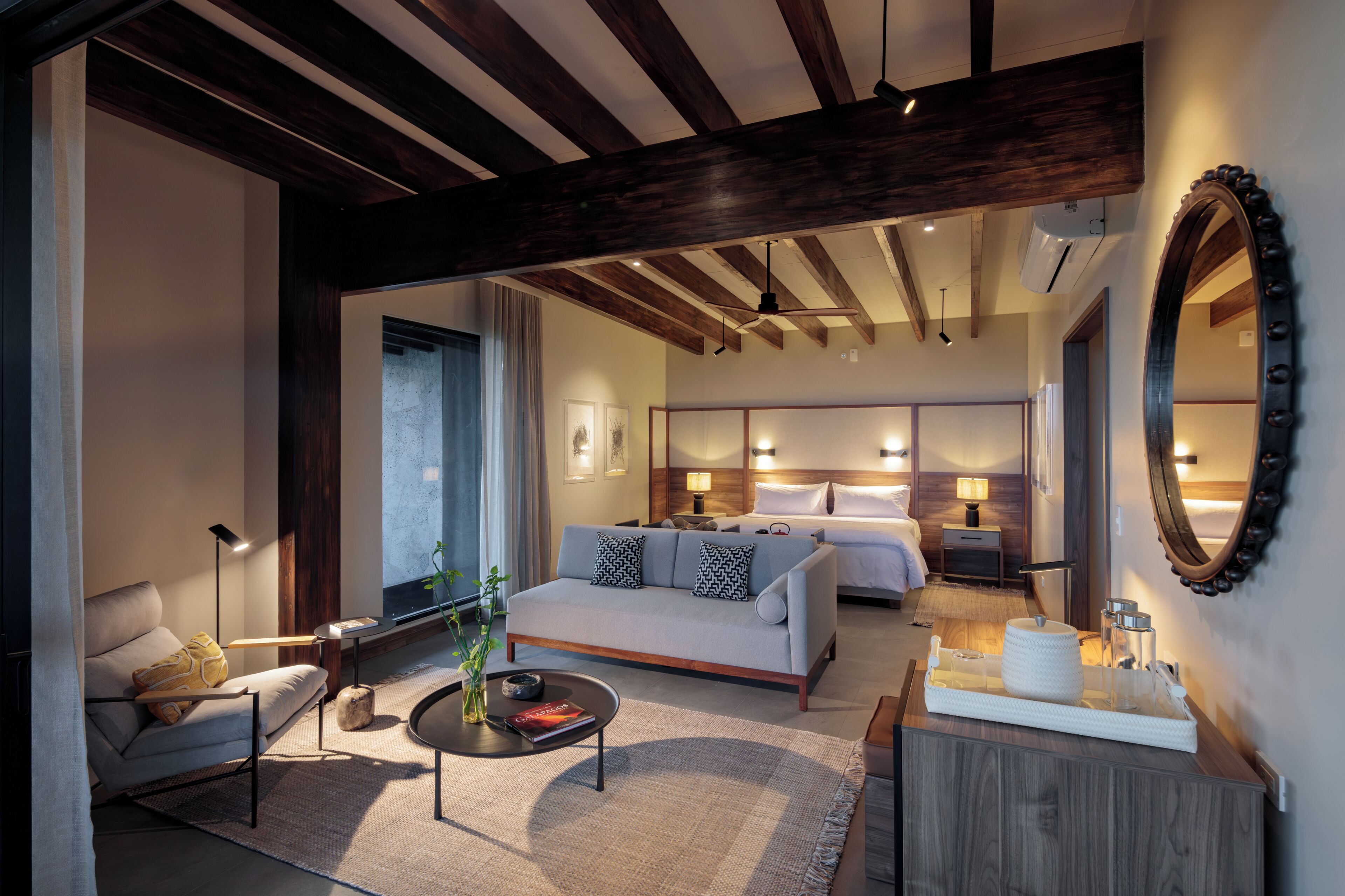 A modern hotel room with a sofa, a bedside table, a lamp, a bed, a mirror, and a large window.