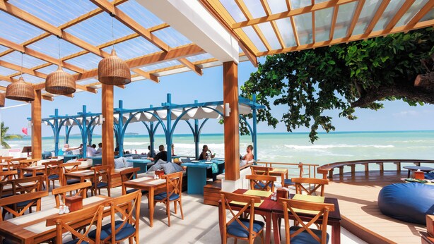 A beachside restaurant with wooden tables and chairs, a covered seating area, and a view of the ocean.