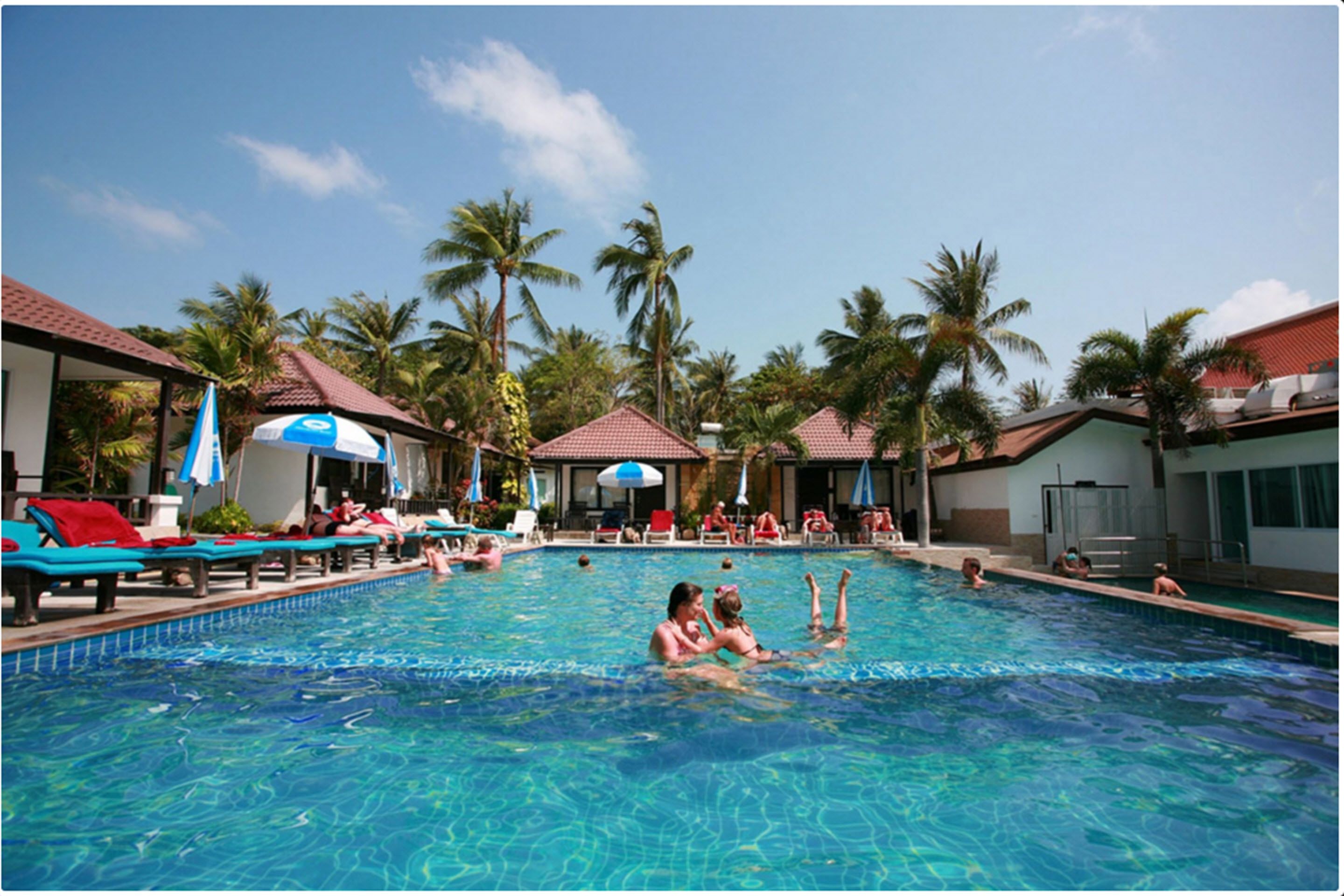 A swimming pool with lounge chairs and umbrellas, surrounded by palm trees and buildings with red roofs.