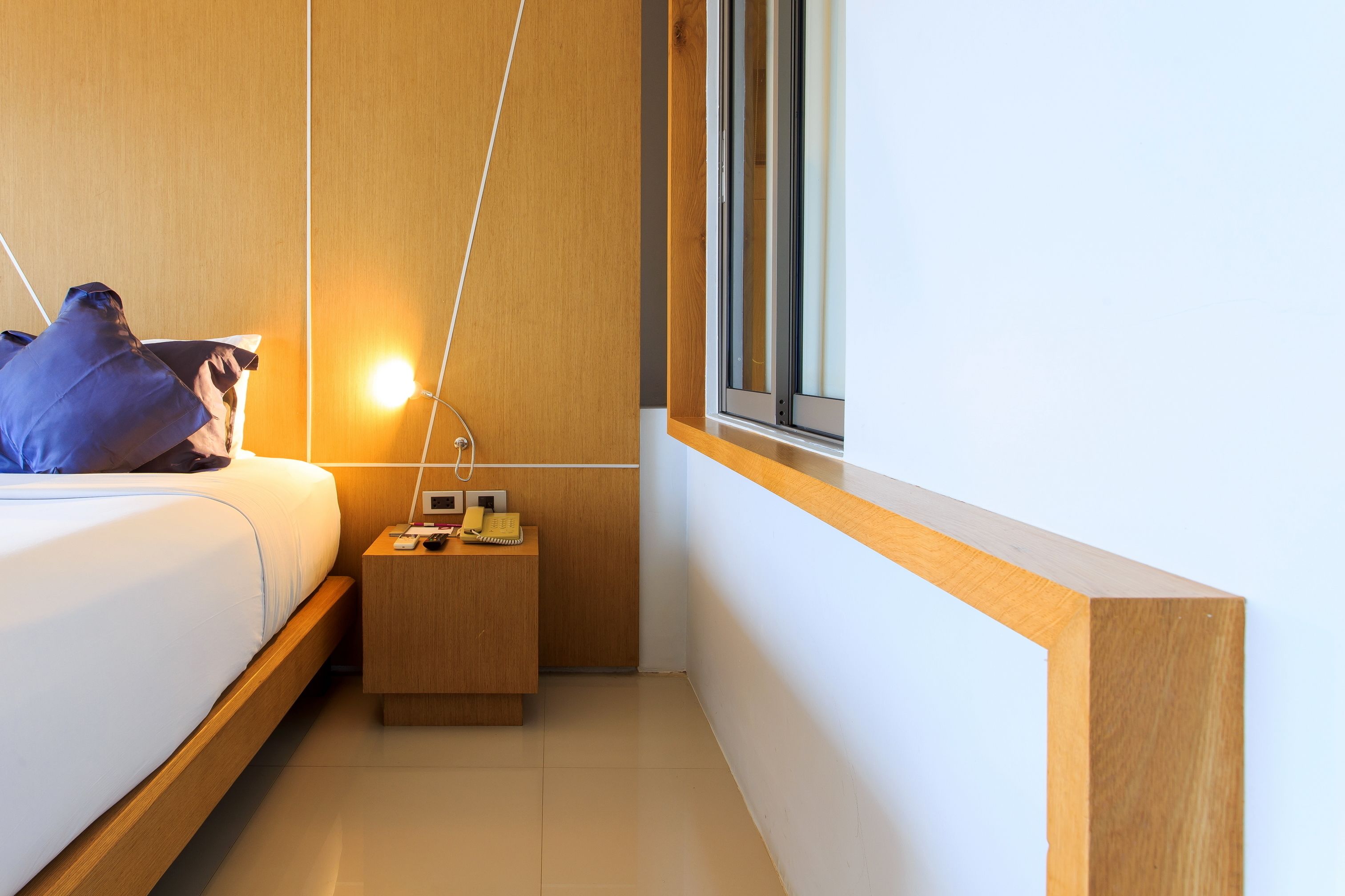 A modern hotel room with a bed, bedside table with a phone, and a window with blinds.