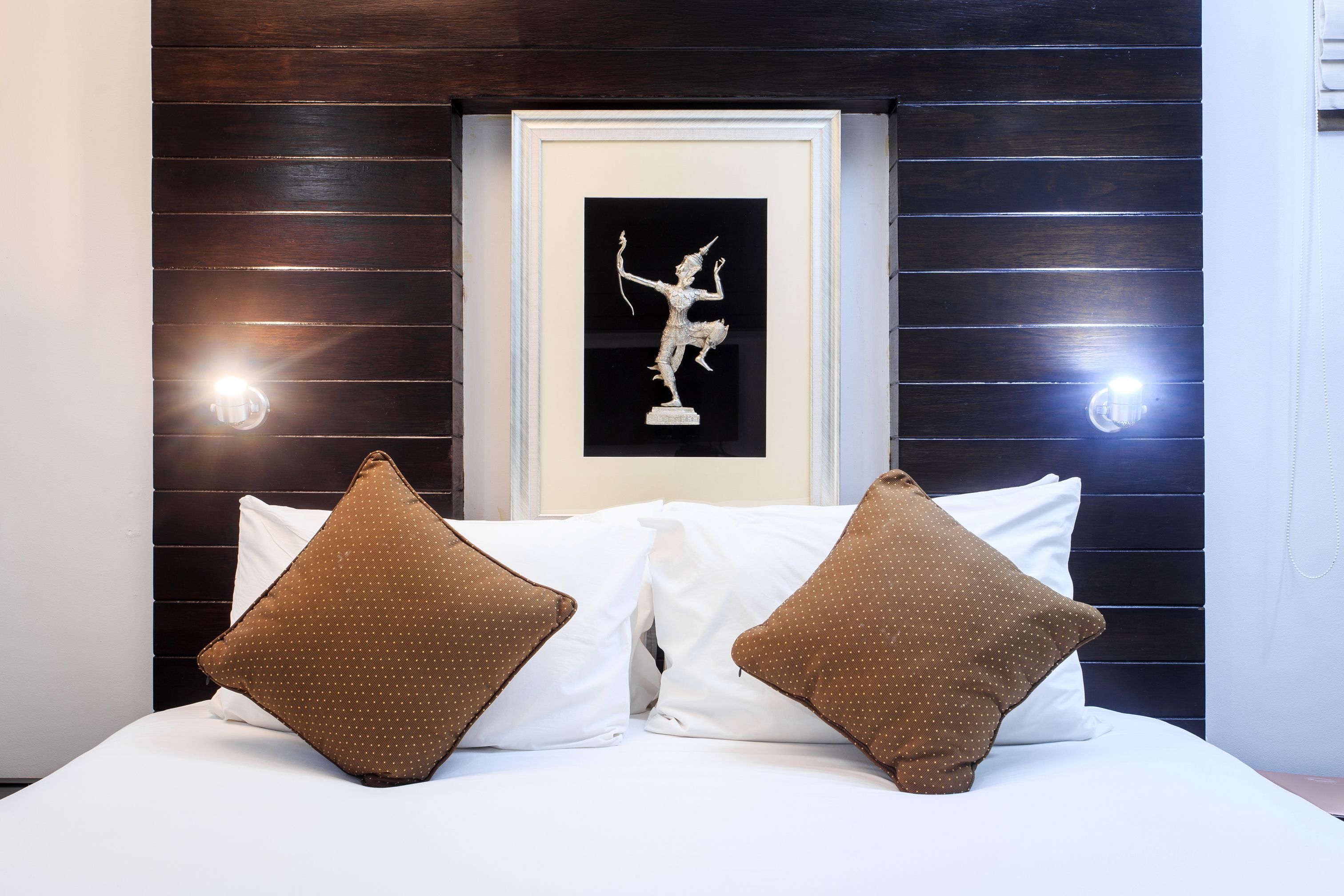 A neatly made bed with white and brown pillows, a framed picture on the wall, and two wall-mounted lights.