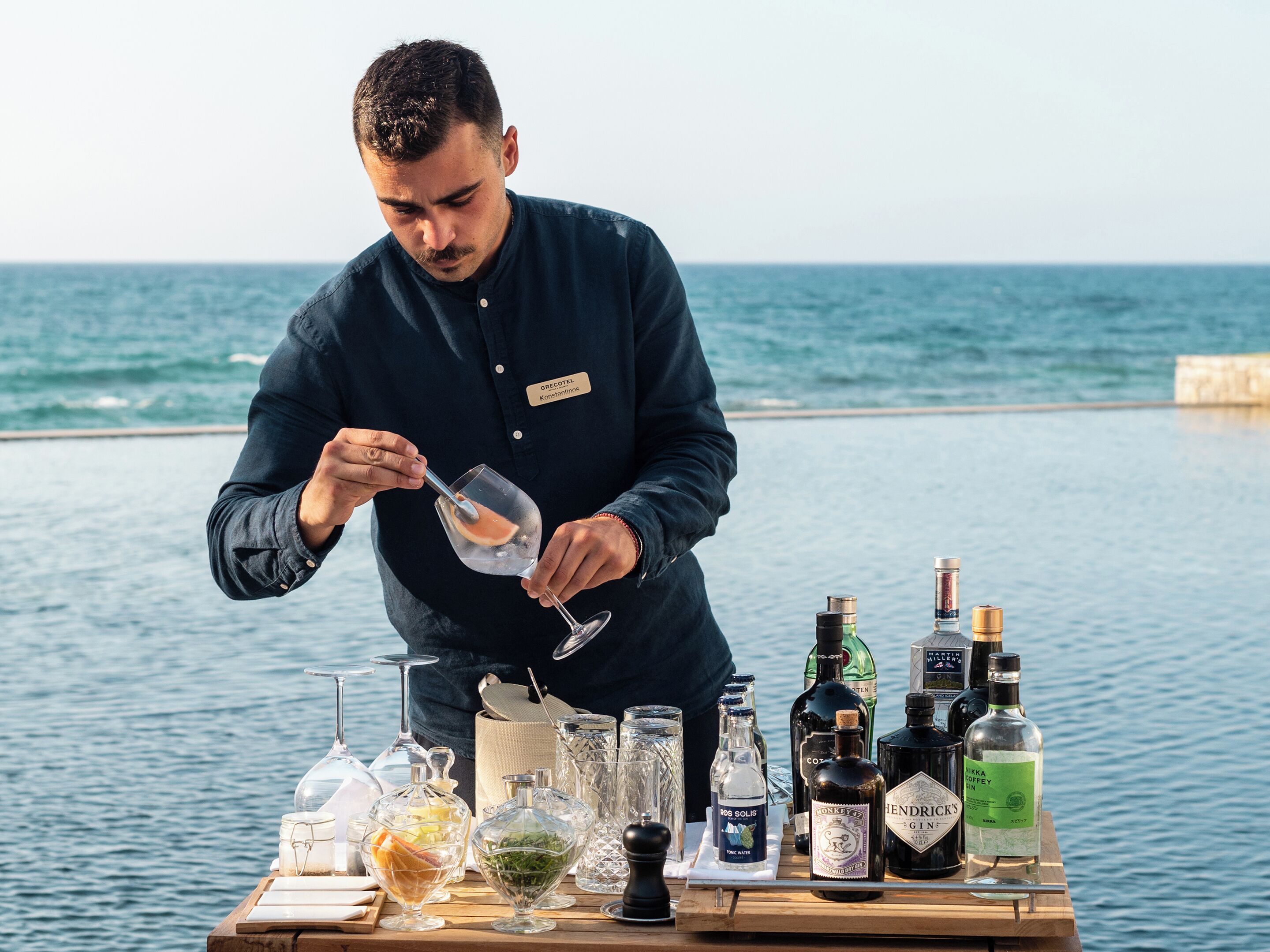 A bartender preparing a drink at a seaside bar with various bottles and glasses on the counter.