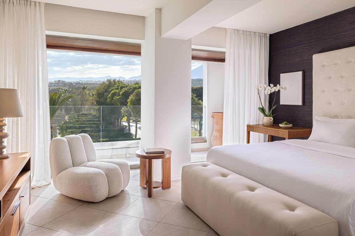 Ein modernes Schlafzimmer mit einem großen Bett, einer Sitzecke, einem kleinen Tisch und Blick auf eine üppige Landschaft.