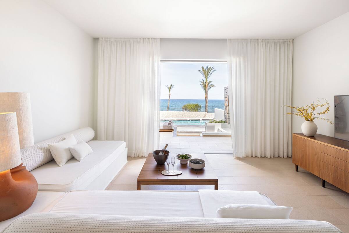 Ein modernes Hotelzimmer mit einem großen Bett, einer Couch, einem Esstisch mit Schüssel und Weingläsern sowie Meerblick und Blick auf Palmen durch das Fenster.