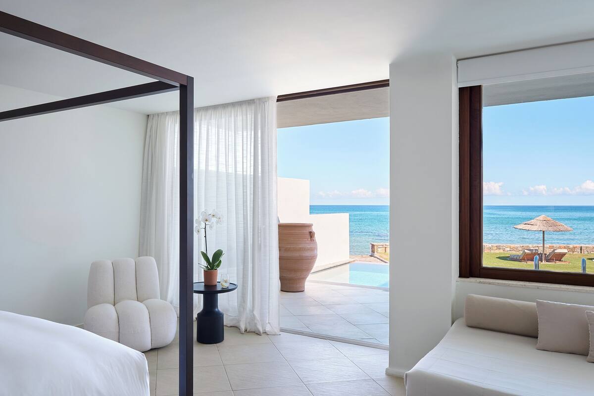 Ein Hotelzimmer mit Bett, einer Sitzecke, einer Topfpflanze, einem großen Fenster mit Meerblick und einem Balkon mit Pool.