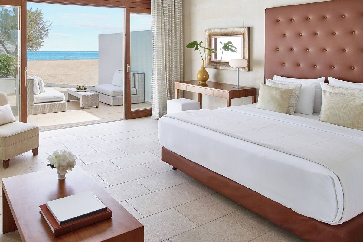 Ein Hotelzimmer mit einem großen Bett, einem Schreibtisch, einem Sessel, einem kleinen Tisch mit einer Vase und Blick auf den Strand.