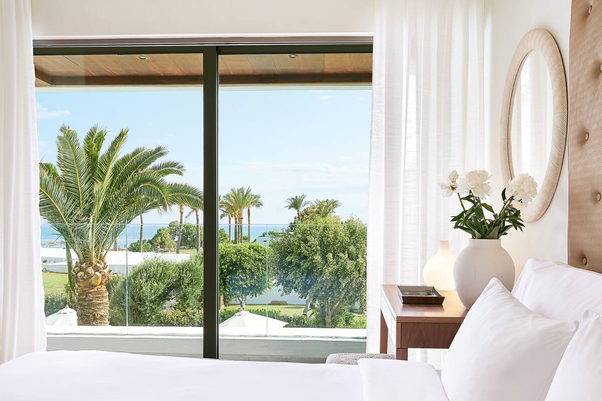 Ein Hotelzimmer mit großem Fenster, einem Bett, einem Nachttisch, einer Vase mit Blumen und einem Spiegel.