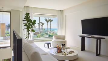 A modern living room with a white sofa, a round coffee table with drinks and snacks, a TV on a stand, and a large window with a view of palm trees and the sea.