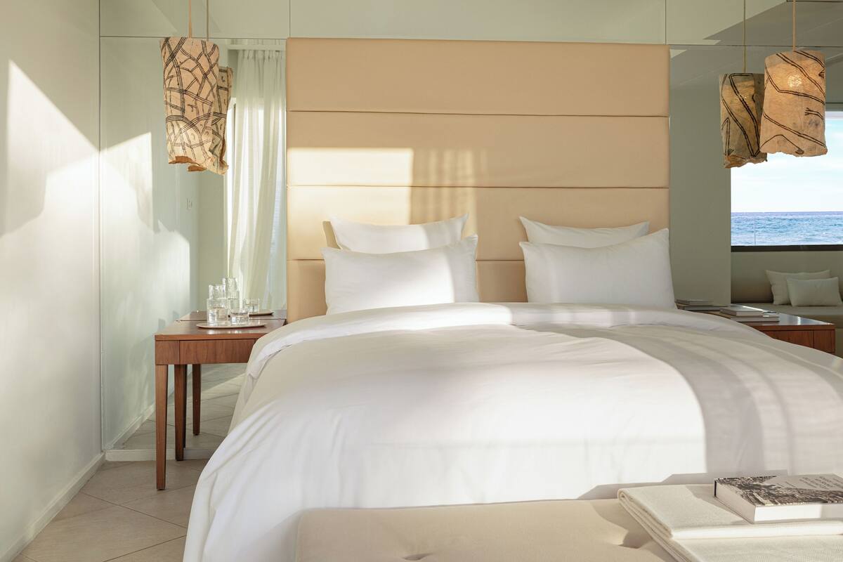 Ein modernes Hotelzimmer mit einem großen Bett, einem Nachttisch mit Gläsern, einem kleinen Tisch mit einem Buch und Blick aufs Meer.