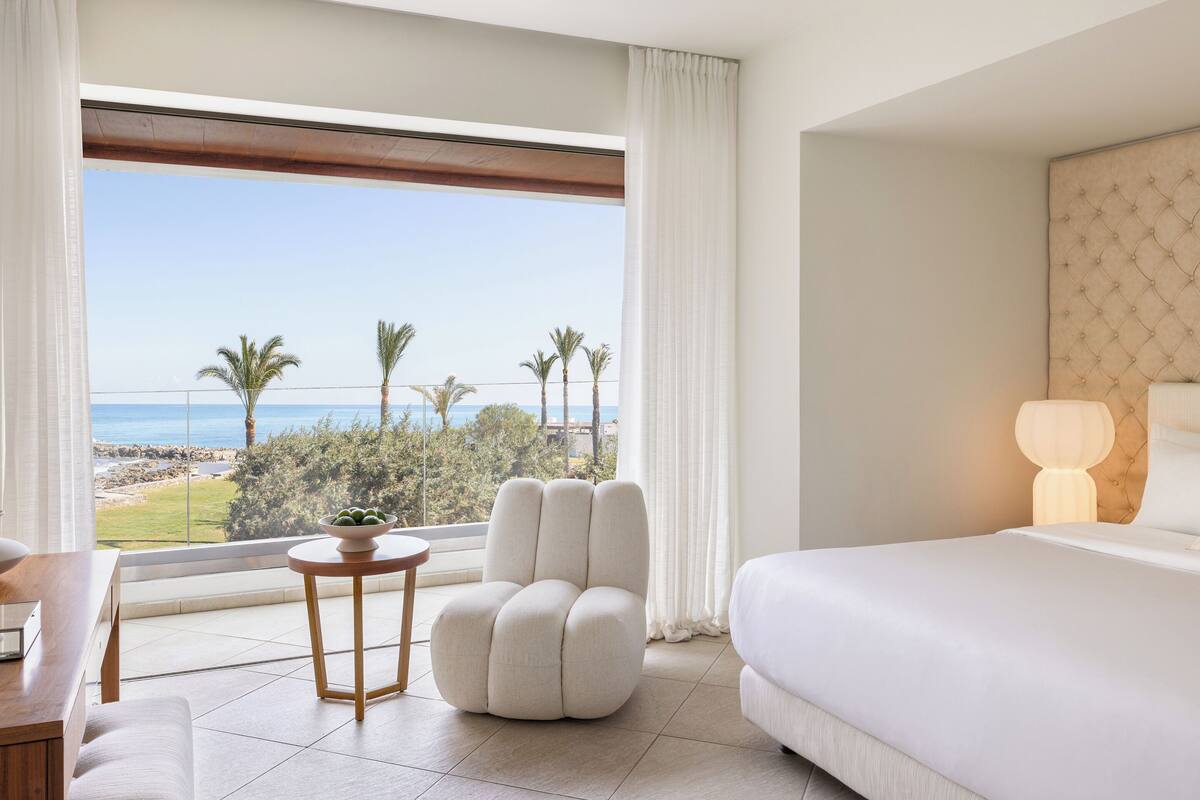 Ein Hotelzimmer mit Bett, einem runden Tisch mit Obstschale, einem Stuhl und Blick auf das Meer und Palmen.