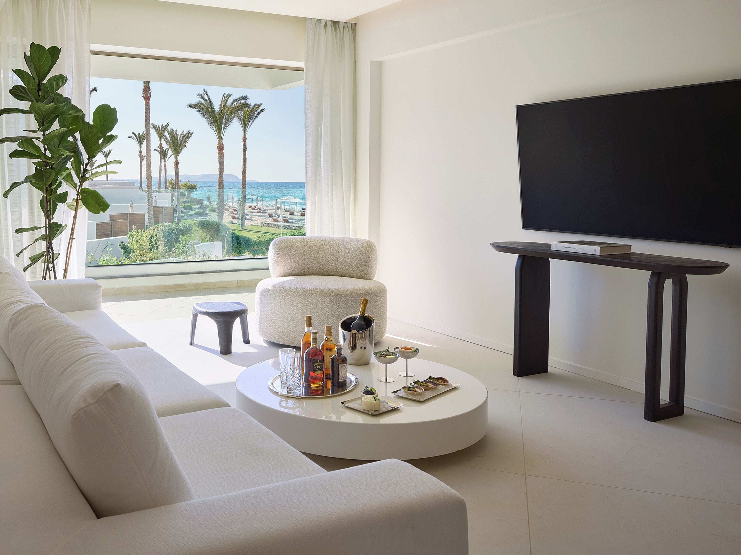 A modern living room with a white sofa, a round coffee table with drinks and snacks, a TV on a stand, and a view of palm trees and the sea through large windows.