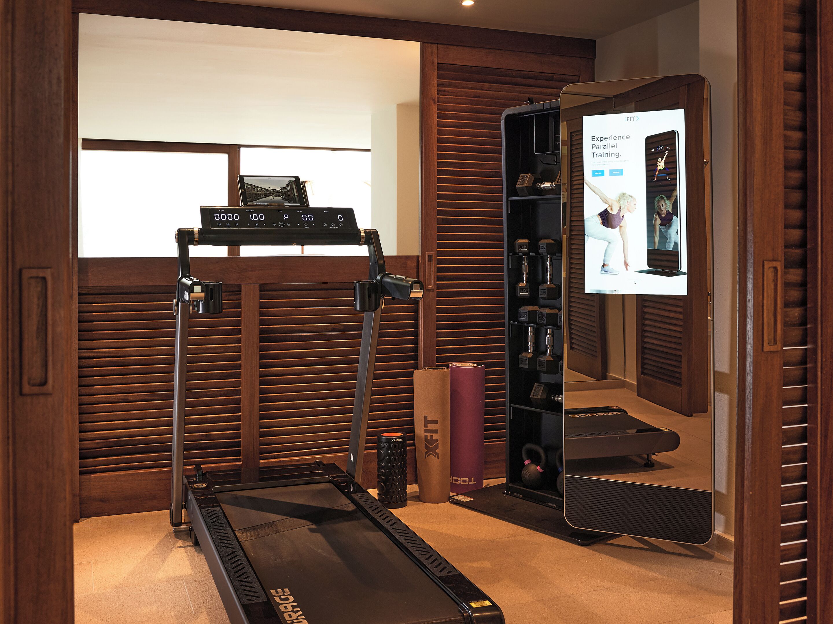 Un gimnasio en casa con cinta de correr, pesas y una televisión mostrando contenido de acondicionamiento físico.
