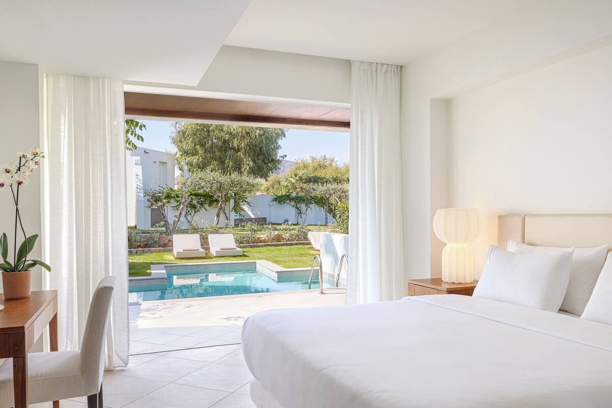 Ein Hotelzimmer mit Bett, Schreibtisch, Stuhl, Lampe und Blick auf Pool und Außenbereich mit Sitzgelegenheiten.