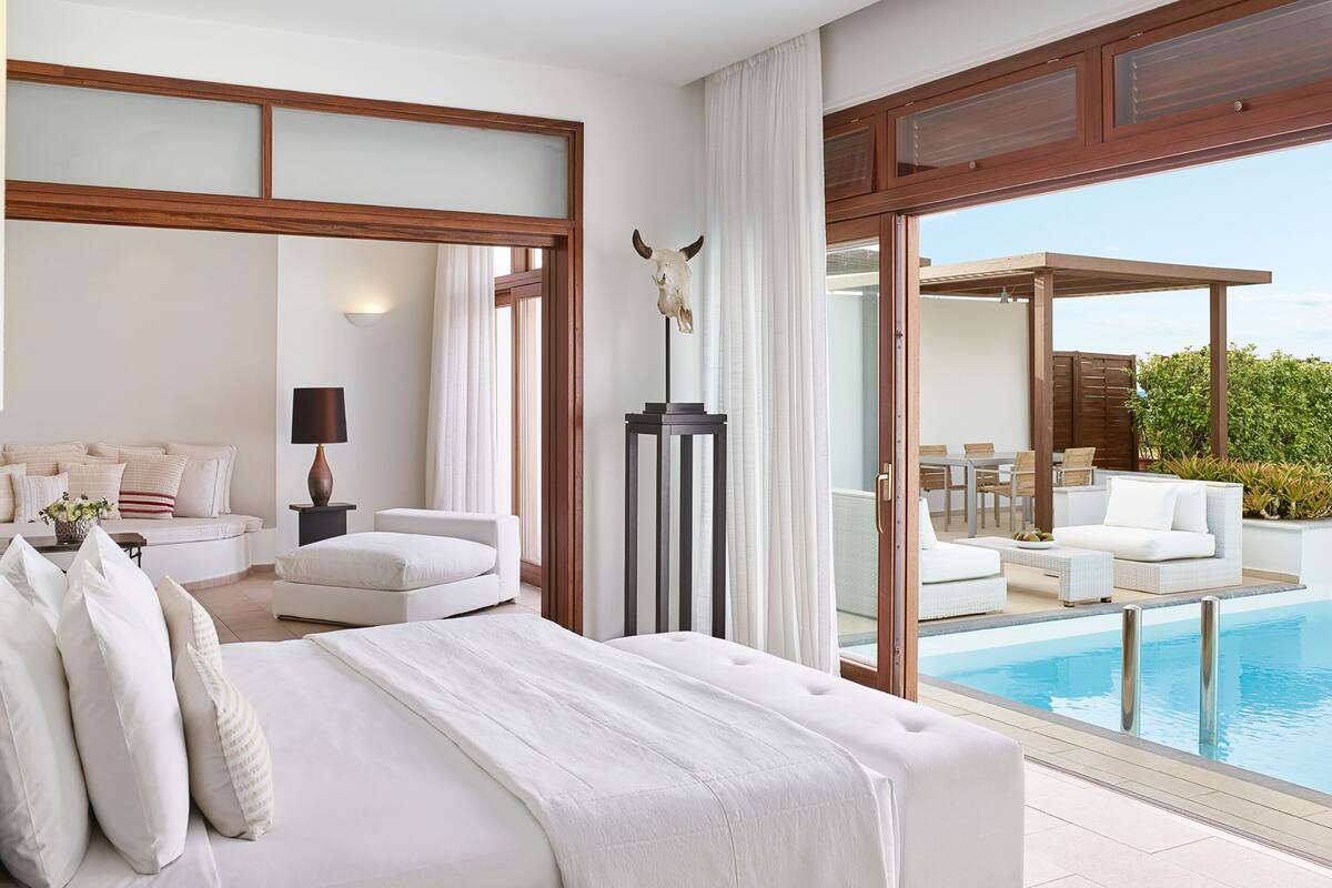 Ein Hotelzimmer mit einem großen Bett, einem Schreibtisch, einem Sessel, einer Lampe, einer Statue und Blick auf einen Außenpool.