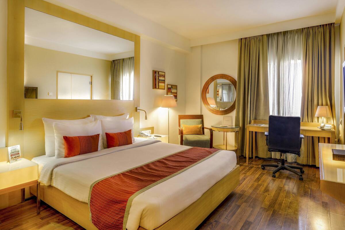 Une chambre d’hôtel avec un grand lit, un bureau, une chaise, un miroir rond et un téléviseur.