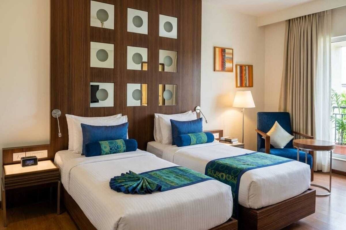Une chambre d’hôtel avec deux lits, une tête de lit en bois, une petite table, une chaise et une fenêtre avec des rideaux.