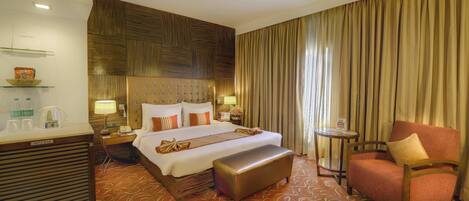 A hotel room with a large bed, a wooden headboard, a bench, a chair, a desk with a telephone, and a small table with a lamp.
