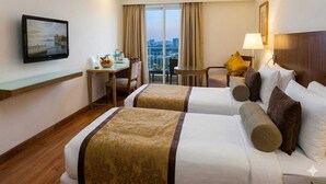 Une chambre d’hôtel avec deux lits, un bureau avec une chaise, une télévision, un balcon avec des rideaux et une lampe.