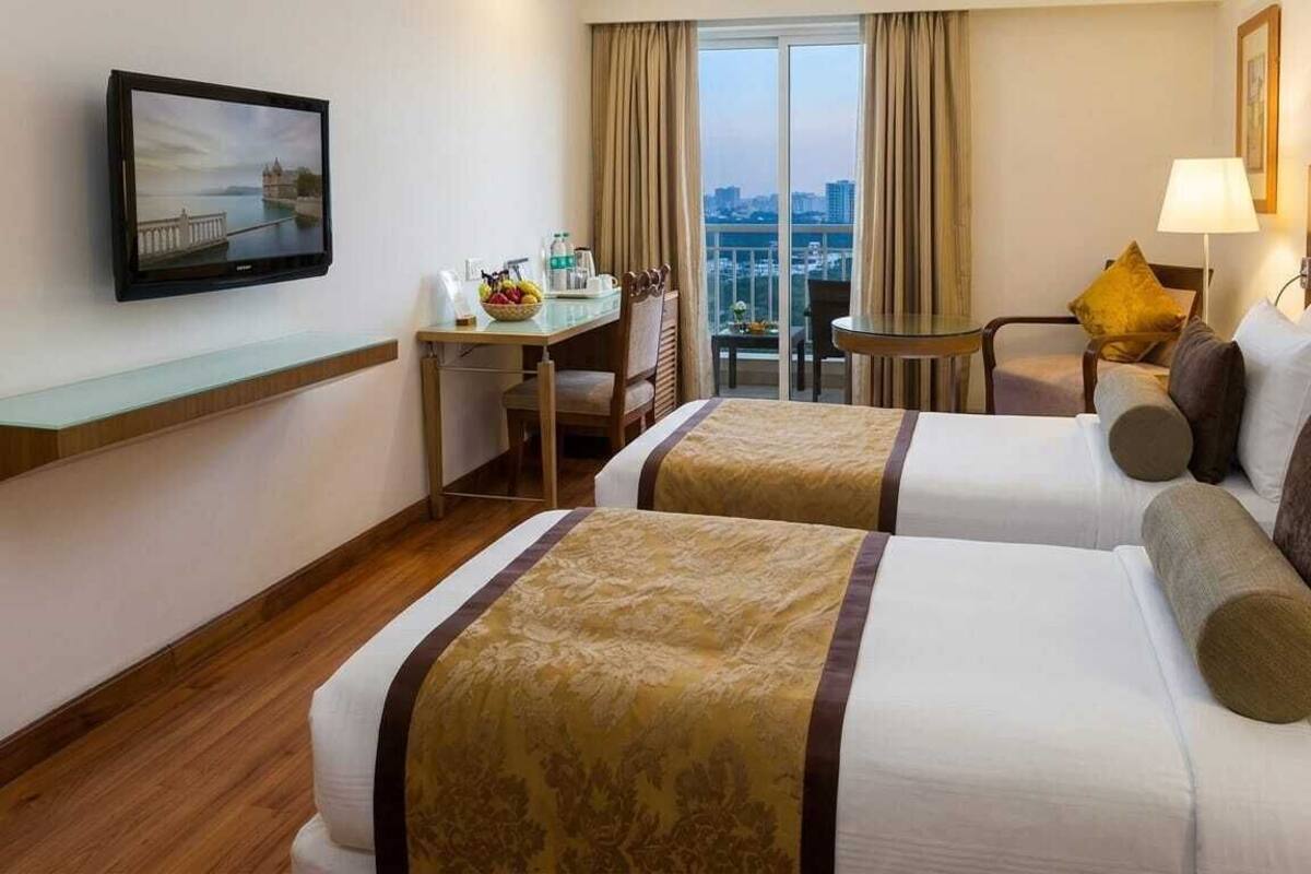 Une chambre d’hôtel avec deux lits, un bureau avec une chaise, une télévision, un balcon avec des rideaux et une lampe.