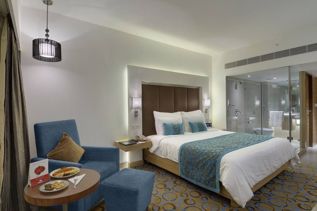 A hotel room with a bed, a desk with a chair, a small table with food, and a shower area.