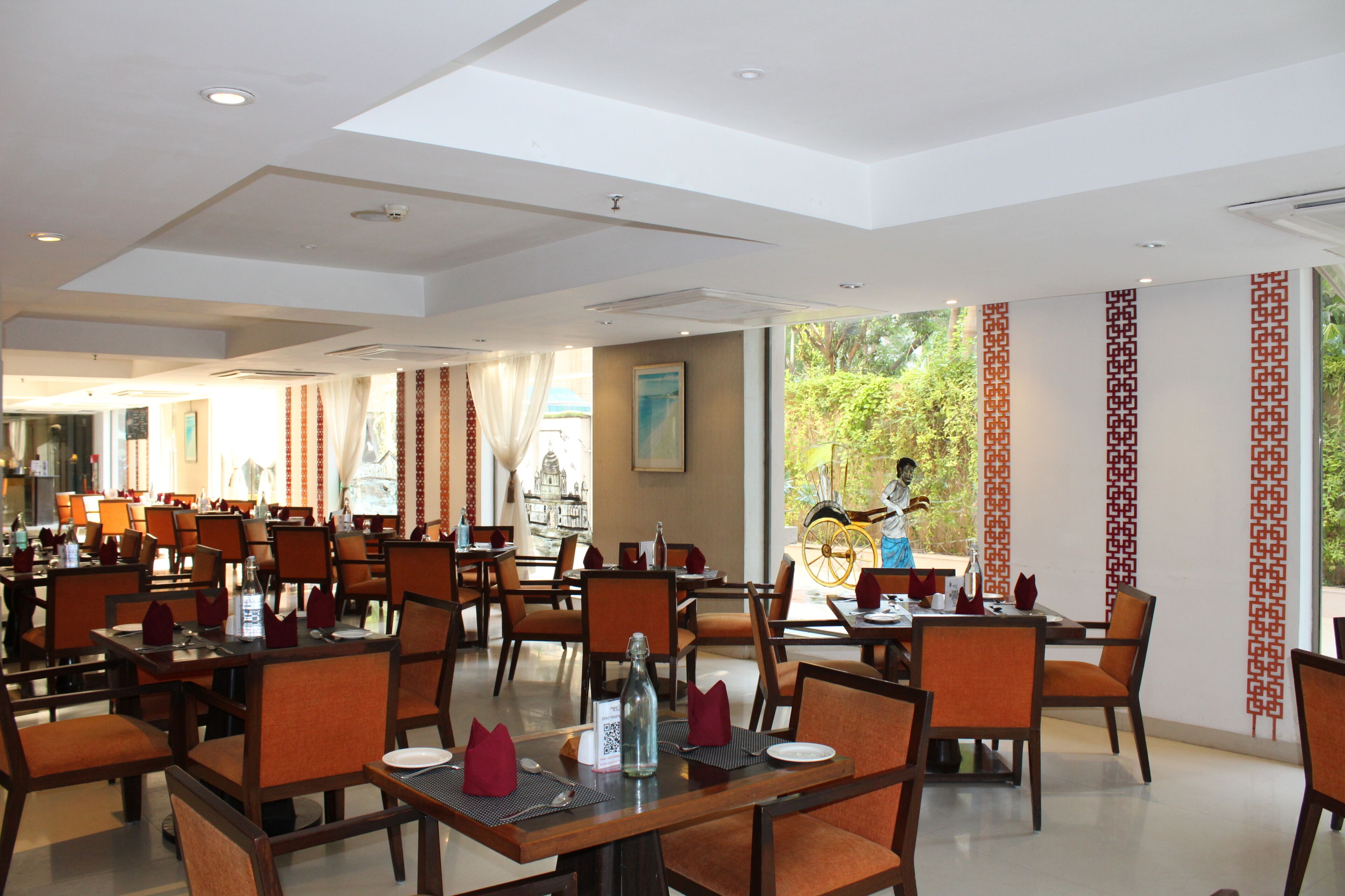 A spacious restaurant with multiple tables and chairs, large windows with a view of greenery, and a high ceiling with skylights.