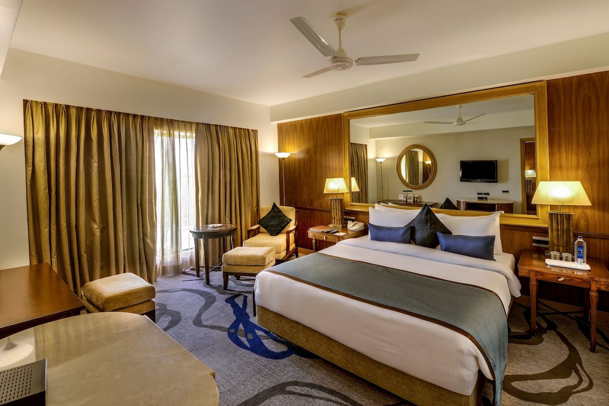 A hotel room with a large bed, a desk, a chair, a desk lamp, a bedside table with a lamp, a mirror, a television, and a ceiling fan.